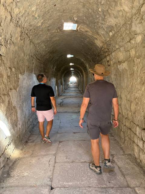 Two travellers walk through a long, stone-arched tunnel lit by daylight at the far end.