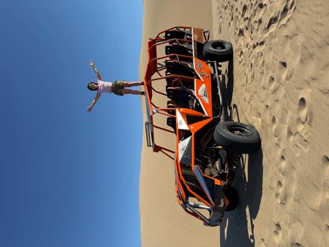 Traveler stands triumphantly on top of an orange dune buggy amid vast golden desert dunes.