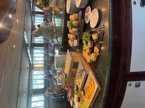Indoor buffet on a cruise ship with assorted cheeses and salads while passengers dine in the background.