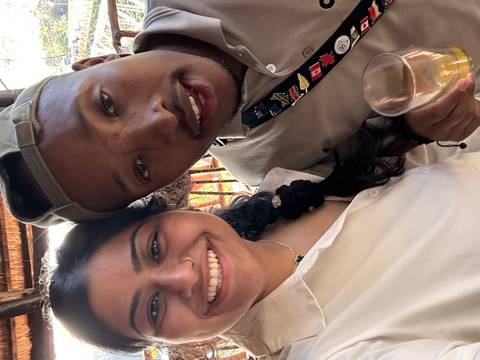 Close-up selfie of two smiling travellers, one holding a drink, inside a wooden venue.