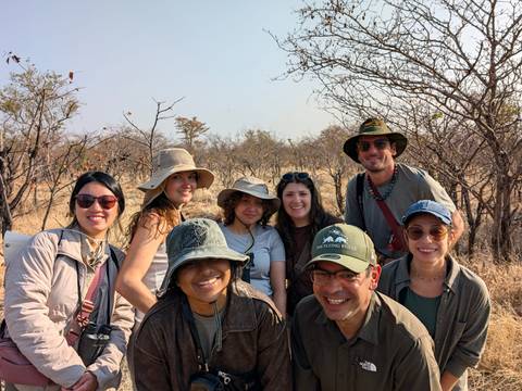 Smiling safari group poses among dry savanna trees under bright midday sun.