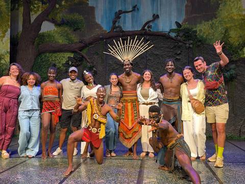 Tourists pose with traditional performers on a stage, vibrant costumes and smiles all around.