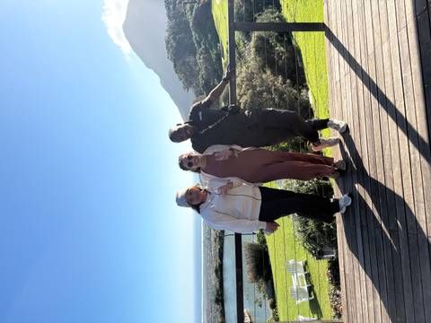 Three friends enjoy wine on a wooden deck overlooking coastal hills and the ocean.