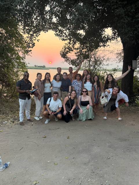 Large tour group poses at sunset by a river with orange sun on horizon.