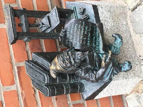 Small bronze dwarf statue sits reading a scroll beside stacked books on a city sidewalk.