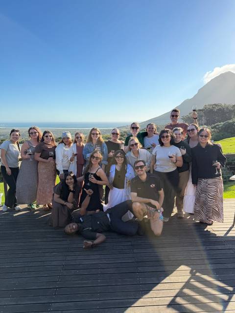 Large group of travelers toasts with wine on a sunny deck overlooking ocean and mountains.