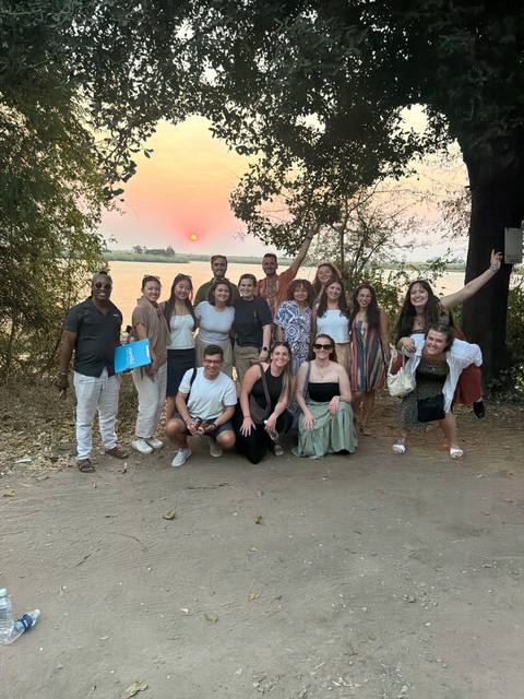 Travel group poses by a riverbank with the sun setting behind distant palms.
