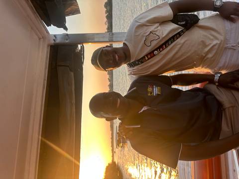 Two men stand on a boat deck at sunset with a tranquil river backdrop.