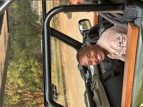 Smiling safari guide sits in the driver seat of an open game-viewing vehicle.