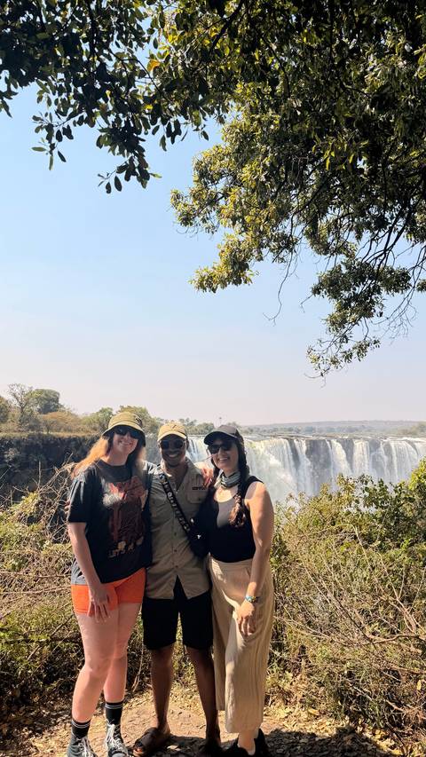 Three travelers pose happily with the misty cascades of Victoria Falls behind them.