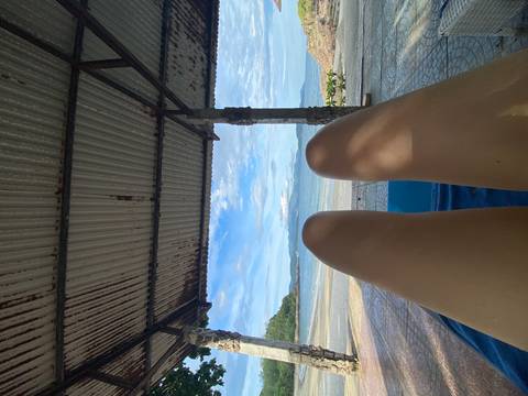 Relaxed viewpoint from under a rustic shelter, legs stretched toward a calm sea horizon.