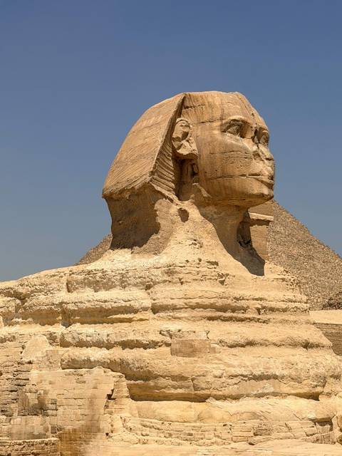 Side profile of the Great Sphinx carved from limestone with a pyramid backdrop under a clear sky.