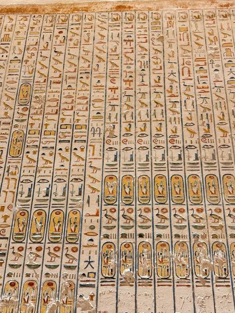 Rows of finely carved hieroglyphic cartouches painted in faded yellows and blues on a temple wall.