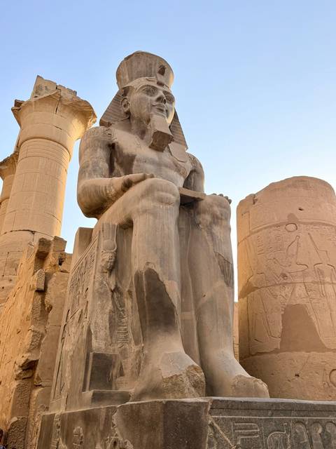 Weather-worn seated pharaoh statue flanked by tall pillars in soft evening light.