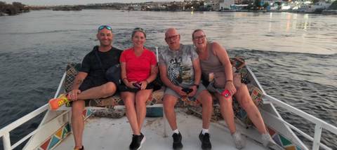 Four travelers sit smiling on the bow of a small boat cruising the Nile at dusk with city lights behind.