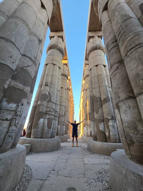 Traveler holds arms wide among towering stone columns of Luxor Temple lit by low sunlight.