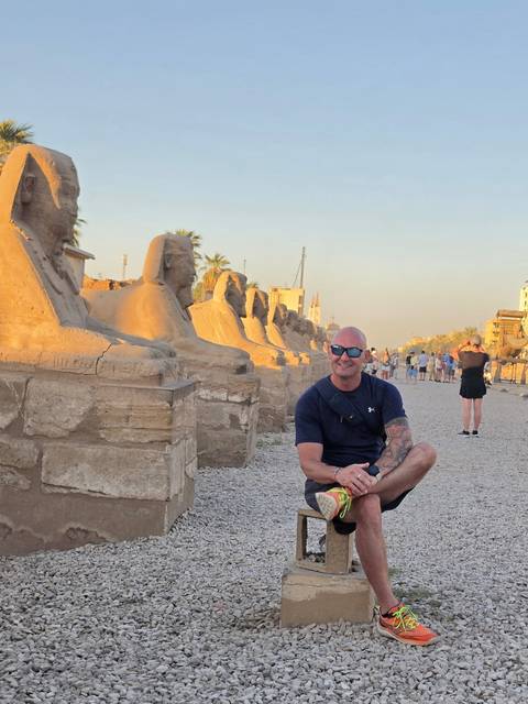 Man kneels for a photo beside a row of ram-headed sphinx statues on the Avenue of Sphinxes.