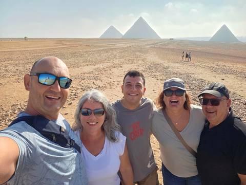 Group selfie in the desert with three pyramids visible on the distant horizon under bright sky.