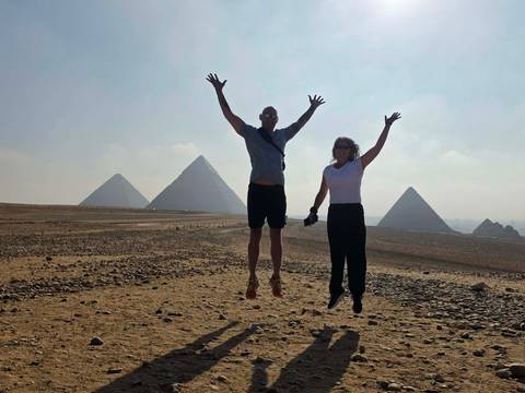 A middle-aged couple jumps joyfully in front of several pyramids on a sunny desert plain.