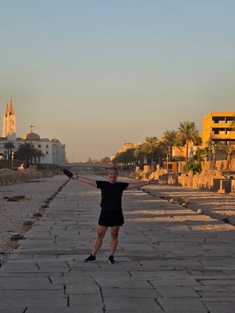 A smiling woman with open arms stands on an ancient stone avenue lined with statues and palm trees during warm sunset light.