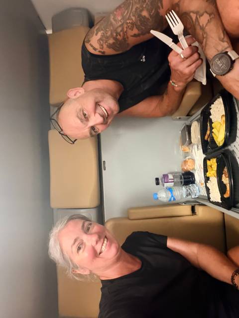 Two travelers enjoy a simple meal in a compact train cabin, smiling toward the camera.