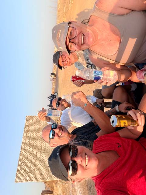 Group of friends raises bottled drinks while sitting on a desert cart road in late afternoon light.