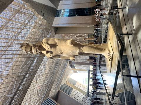 Colossal pharaoh statue stands beneath a dramatic glass ceiling inside a modern museum atrium.
