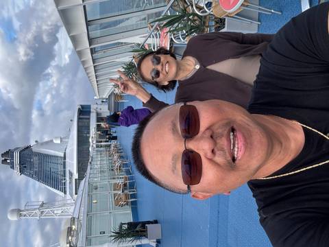 Cheerful couple takes a selfie on the open deck of a large cruise ship under partly cloudy skies.