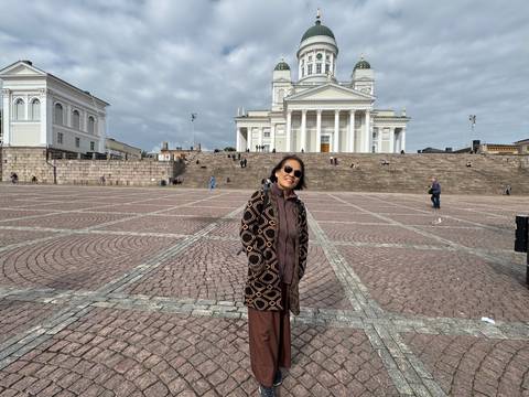 Woman poses in wide cobblestone square with Helsinki Cathedral in background under cloudy sky.