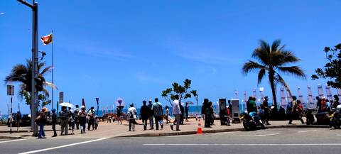 Busy seaside promenade with crowds, a Sri Lankan flag, palm trees and blue ocean beyond.