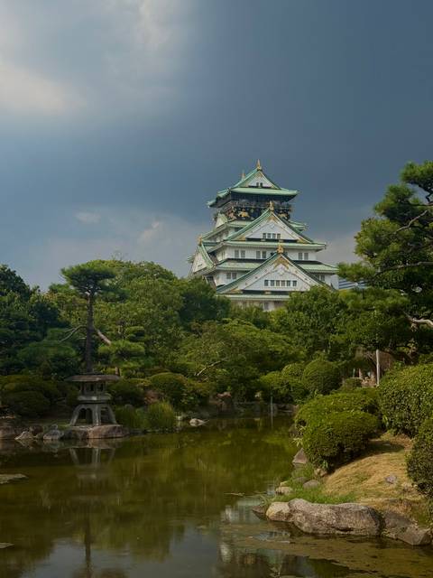 Osaka Castle’s green-tiled roofs rise above lush trees under a moody sky