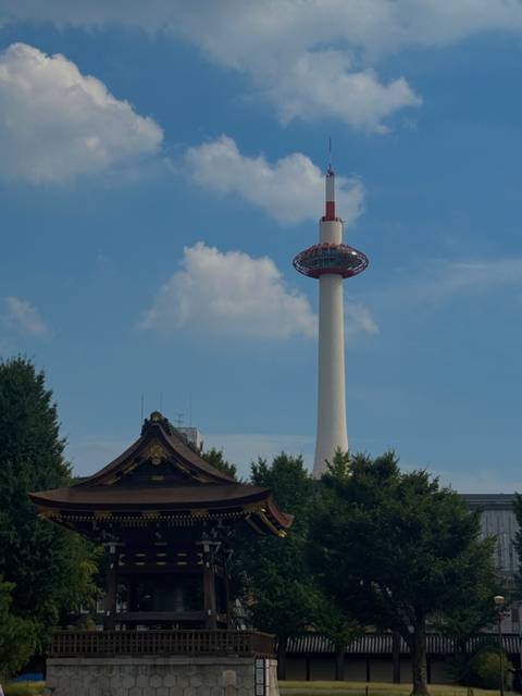 Kyoto Tower rises behind the ornate roof of a traditional temple under a blue sky with clouds