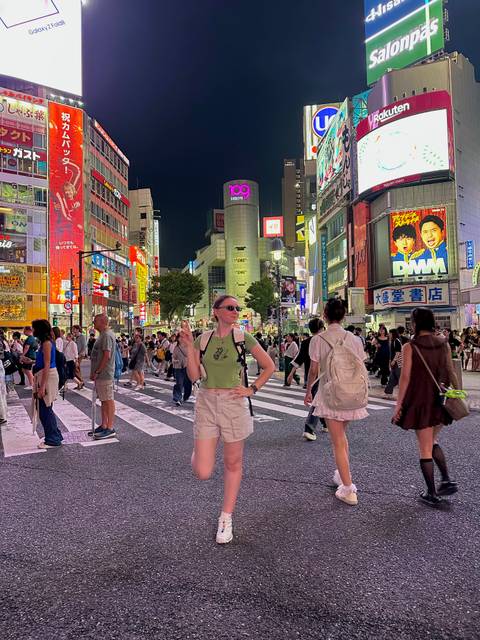 Vibrant night scene at Shibuya Crossing with crowds, neon billboards and a traveler posing in the foreground