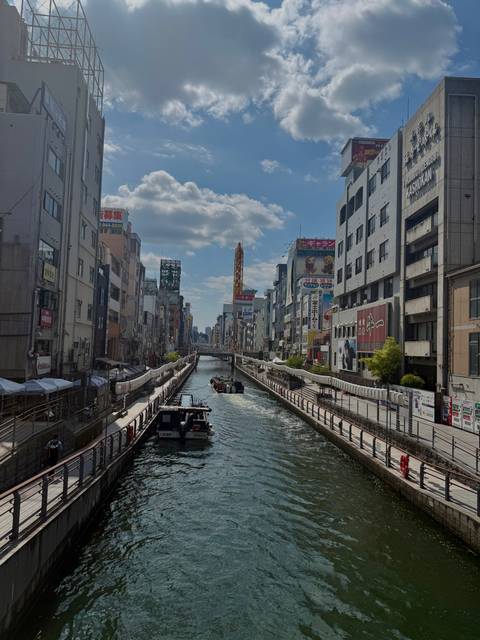 Daytime scene along Osaka’s Dotonbori canal lined with tall commercial buildings and billboards.