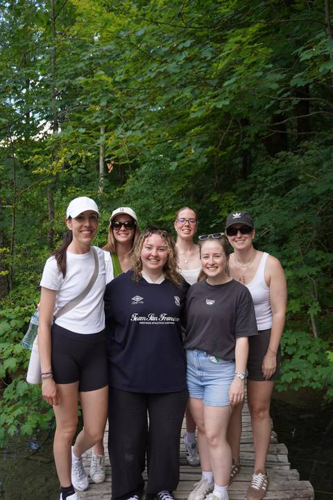 Six travelers stand together on a forest trail surrounded by dense green foliage.