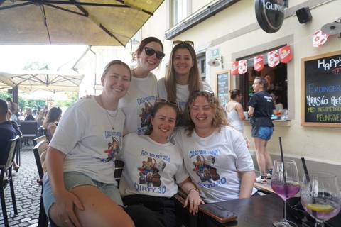 Five friends wearing matching birthday shirts pose at a street-side café.