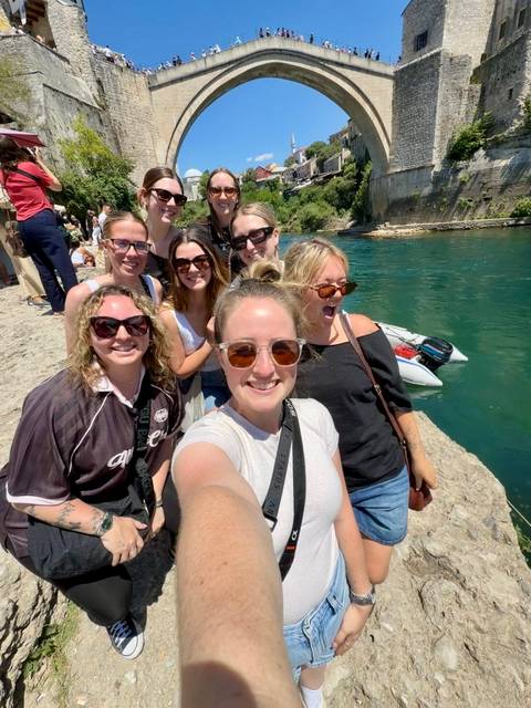 A cheerful group of young travellers taking a selfie on a stone riverside path beside clear turquoise water.