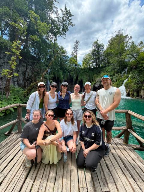 Travellers posing on a wooden bridge with emerald pools, lush forest and cascading waterfalls behind them.