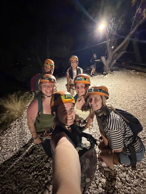 Group wearing helmets and harnesses posing on a gravel trail at night before an adventure activity.