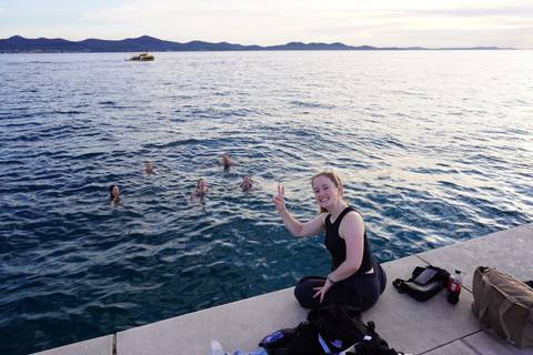 Young woman flashing a peace sign on a concrete pier while friends swim in a calm sea at sunset.