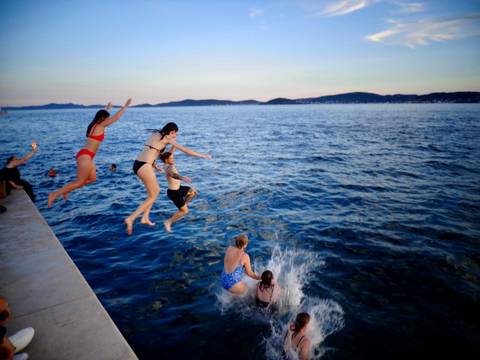Thrilled travellers mid-air as they leap from a pier into the Adriatic Sea at dusk.