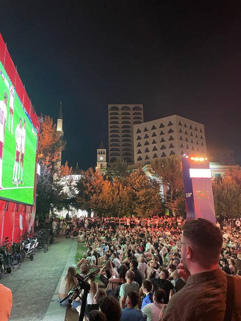 Nighttime public viewing area with a giant outdoor screen showing football in a city square.