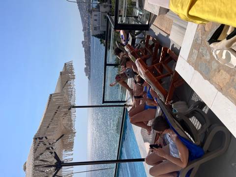 Travellers sunbathing on pool loungers overlooking a sparkling bay and coastal town.