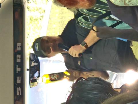 Tour guide on a coach enthusiastically holds up a bottle and speaks into a microphone to passengers.