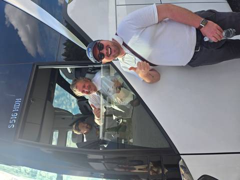 Man gives a thumbs-up outside a modern tour bus while driver and another passenger smile from inside the cab.
