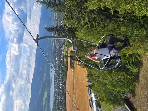 Traveller rides a chairlift high above farmland and a lake while filming the view with a phone.