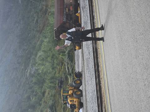 Railway staff member in uniform waves from beside the tracks with heavy machinery in the background.