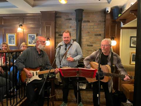 Two guitarists and a singer perform live folk music in a cosy Irish pub while guests look on.