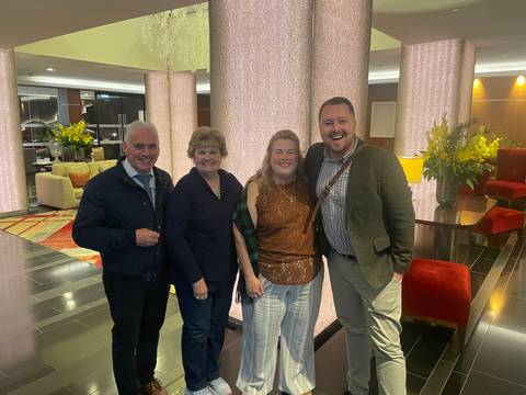 Four travellers pose together in a hotel lobby decorated with tall illuminated columns and fresh flowers.