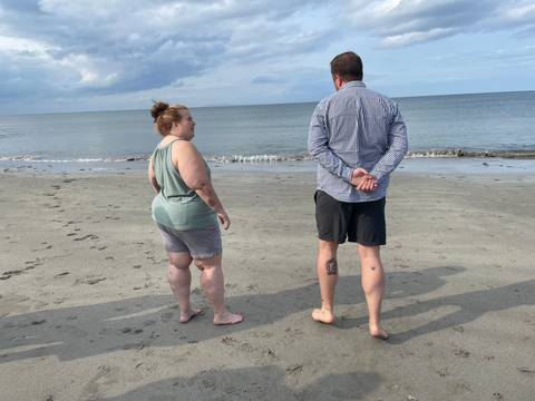 Two people stand barefoot on a quiet grey sand beach looking out over calm sea and sky.